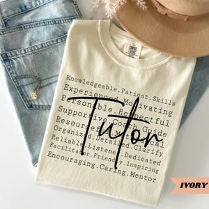 May include: Ivory-colored t-shirt with the word "Tutor" in a bold, black script. The shirt features a list of qualities such as "Knowledgeable," "Patient," and "Supportive." The shirt is displayed with blue jeans and a hat.