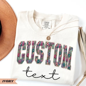 May include: Ivory-colored t-shirt with the word "CUSTOM" in large, floral-filled letters and the word "text" in a handwritten font. A gold necklace and a brown hat are also visible.