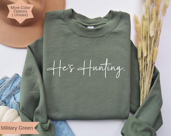 May include: Military green unisex sweatshirt with the text "He's Hunting" in white script. The sweatshirt is folded, with a hat and jeans in the background. The sweatshirt is a crew neck style.