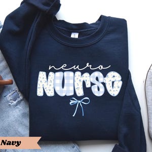 May include: Navy blue sweatshirt with the words "neuro nurse" in white script and block letters. The word "nurse" has a blue gingham pattern with small floral accents. A light blue bow is below the word "nurse".