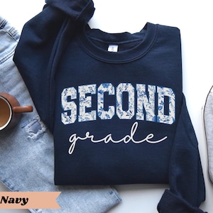 May include: Navy blue sweatshirt with a white floral design that says "Second Grade".