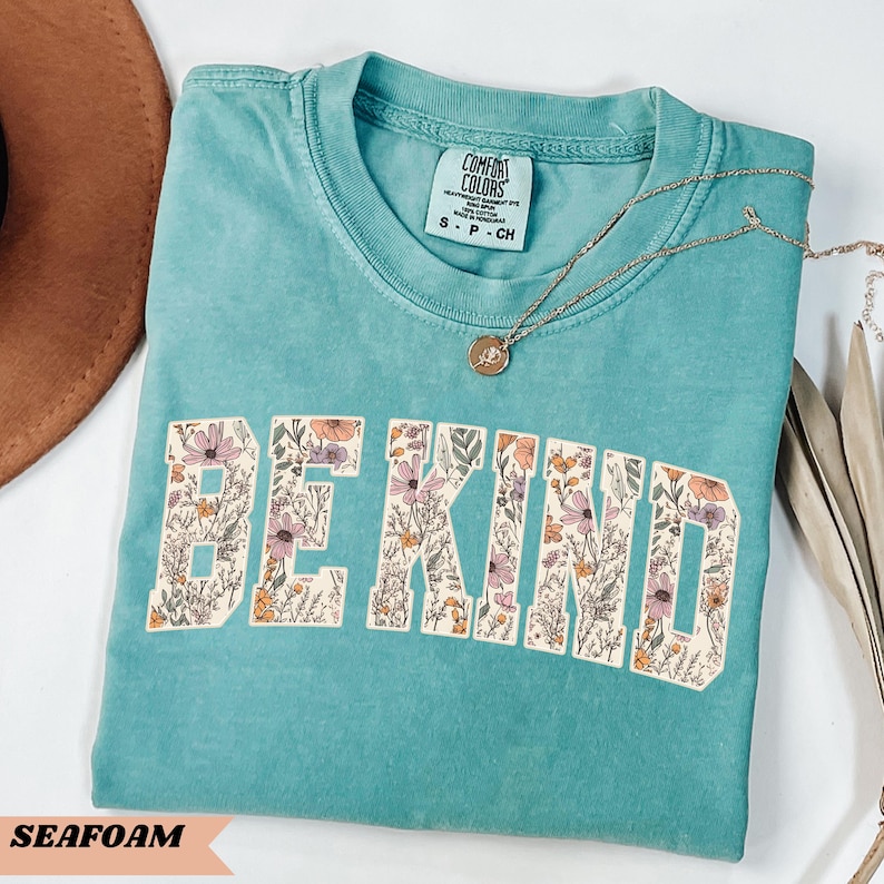 May include: Seafoam green t-shirt with the words "BE KIND" in a floral design. The shirt has a crew neck and short sleeves. A gold necklace with a pendant is draped across the shirt. The shirt is labeled "Comfort Colors".