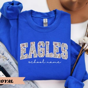 May include: Royal blue sweatshirt with the word "EAGLES" in a floral design. The words "school name" are written in a script font below the word "EAGLES". The sweatshirt is folded with the sleeves on top.