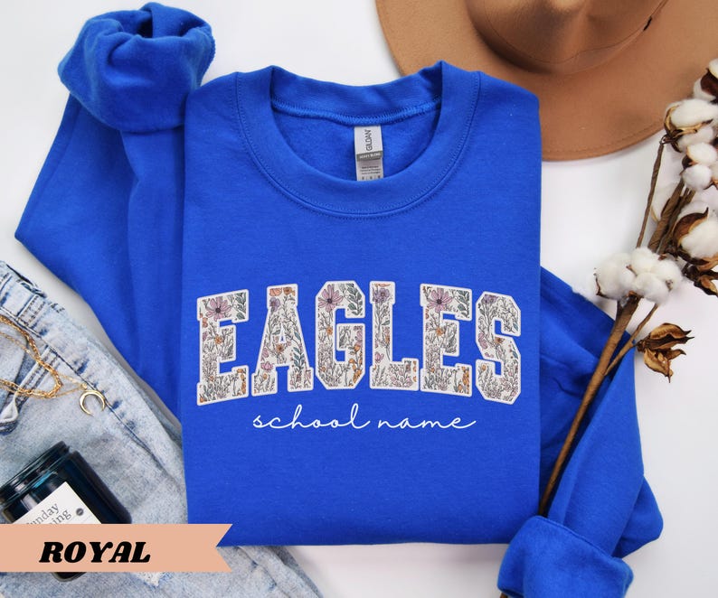 May include: Royal blue sweatshirt with the word "EAGLES" in a floral design. The words "school name" are written in a script font below the word "EAGLES". The sweatshirt is folded with the sleeves on top.