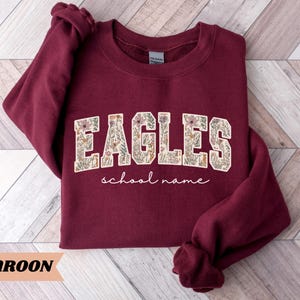 May include: Maroon sweatshirt with the word "EAGLES" in a floral design, with the words "school name" underneath. The sweatshirt is a solid maroon color. The word "MAROON" is written in a tan banner at the bottom.