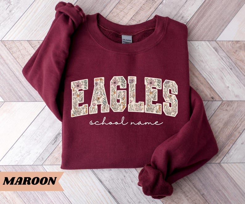 May include: Maroon sweatshirt with the word "EAGLES" in a floral design, with the words "school name" underneath. The sweatshirt is a solid maroon color. The word "MAROON" is written in a tan banner at the bottom.