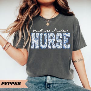 Puede incluir: Una camiseta gris con el texto "neuro NURSE" en letras blancas con un patr&oacute;n floral azul. El texto est&aacute; en una fuente en negrita, de bloque.