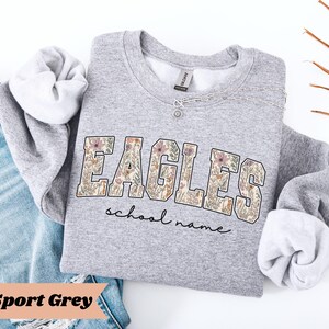 May include: A light gray sweatshirt with the word "EAGLES" in a floral design. Below the word "EAGLES" is the text "school name" in a script font. The sweatshirt is folded with the sleeves rolled up, and is next to a pair of blue jeans.