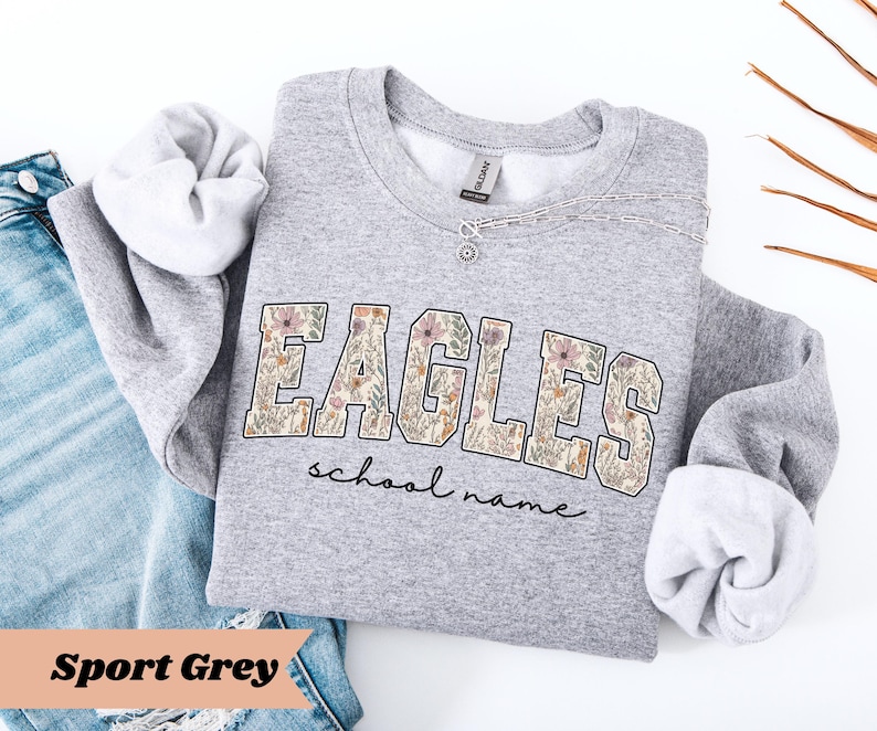 May include: A light gray sweatshirt with the word "EAGLES" in a floral design. Below the word "EAGLES" is the text "school name" in a script font. The sweatshirt is folded with the sleeves rolled up, and is next to a pair of blue jeans.