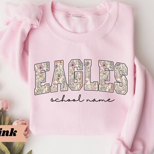 May include: A light pink sweatshirt with the word "EAGLES" in a floral design. Below the word "EAGLES" is the phrase "school name" in a script font. The sweatshirt is folded, with the sleeves visible.