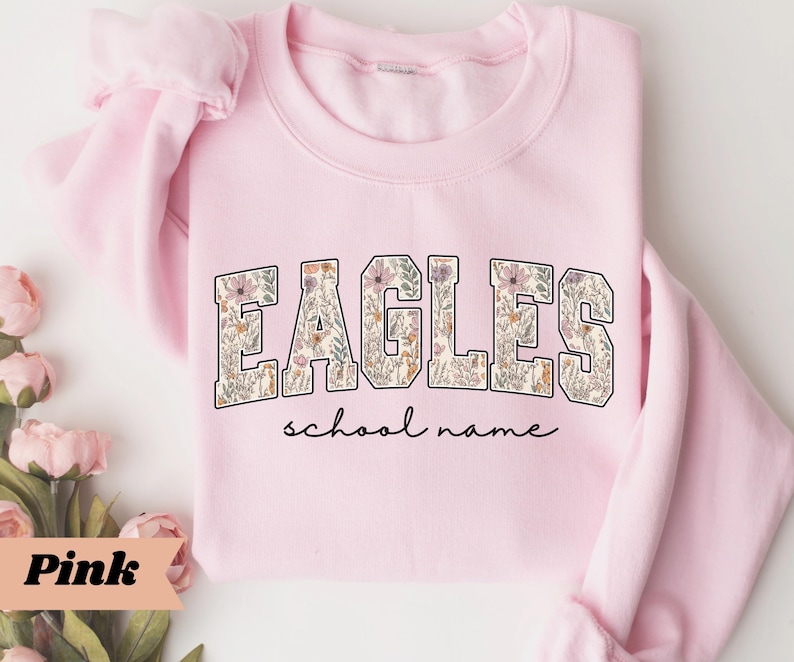 May include: A light pink sweatshirt with the word "EAGLES" in a floral design. Below the word "EAGLES" is the phrase "school name" in a script font. The sweatshirt is folded, with the sleeves visible.