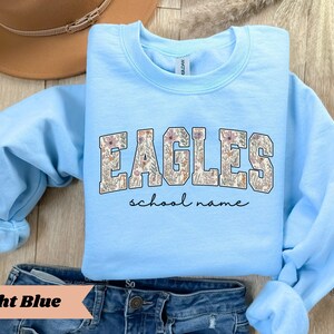 May include: Light blue sweatshirt with the word "EAGLES" in a floral design. Below the word "EAGLES" is the phrase "school name" in a handwritten font. The sweatshirt is displayed on top of blue jeans.