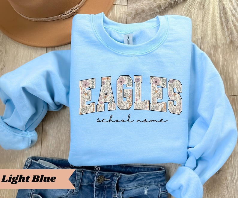 May include: Light blue sweatshirt with the word "EAGLES" in a floral design. Below the word "EAGLES" is the phrase "school name" in a handwritten font. The sweatshirt is displayed on top of blue jeans.