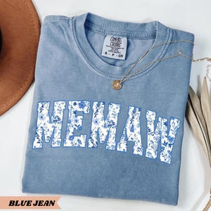 May include: A blue jean t-shirt with the word "MEMAW" printed in white with a floral pattern. The shirt is made of a soft, comfortable material.