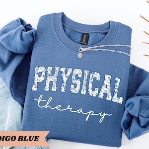 May include: Indigo blue sweatshirt with the words "PHYSICAL THERAPY" in white, with a floral design in the letters. The words "therapy" are written in a cursive font. A silver necklace with a pendant is on the sweatshirt.