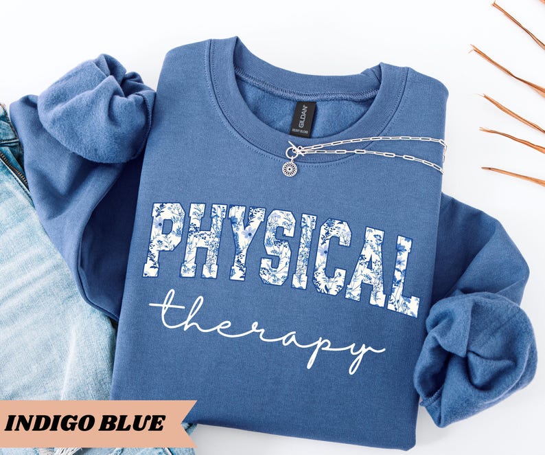 May include: Indigo blue sweatshirt with the words "PHYSICAL THERAPY" in white, with a floral design in the letters. The words "therapy" are written in a cursive font. A silver necklace with a pendant is on the sweatshirt.