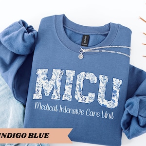 May include: Indigo blue crewneck sweatshirt with the letters MICU in a floral design. Below the letters is the text "Medical Intensive Care Unit". A silver necklace with a pendant is draped across the sweatshirt. The sweatshirt is folded next to a pair of blue jeans.