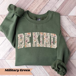 May include: A military green sweatshirt with the words "BE KIND" in a floral design across the chest. The sweatshirt has long sleeves and a crew neck. The bottom of the image has the words "Military Green".