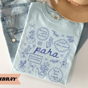 May include: A light blue t-shirt featuring dark blue line art illustrations of flowers, bows, a teapot, and a watering can. The text includes "para", "learning with kindness", "watch them grow", "planting seeds of knowledge", and "teach love inspire".