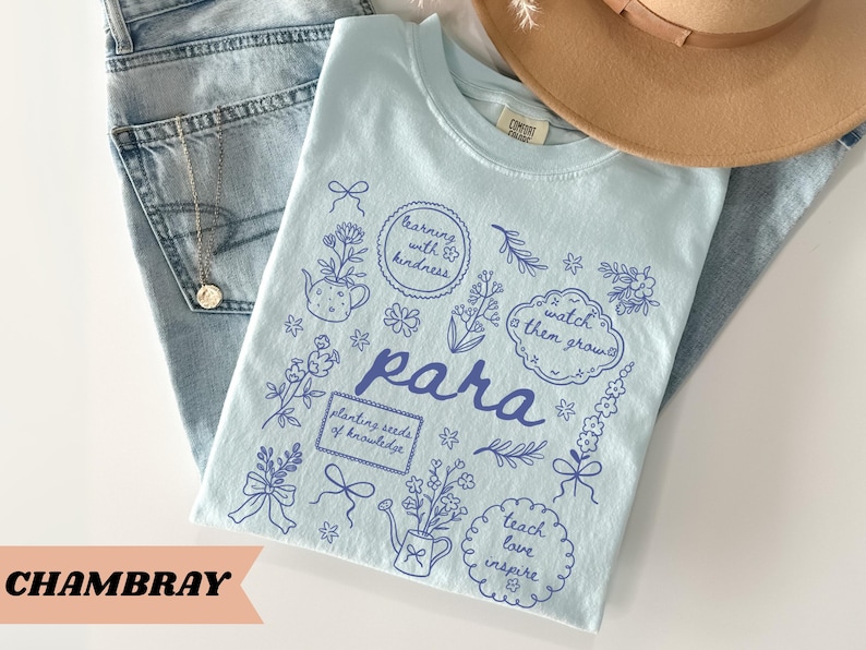 May include: A light blue t-shirt featuring dark blue line art illustrations of flowers, bows, a teapot, and a watering can. The text includes "para", "learning with kindness", "watch them grow", "planting seeds of knowledge", and "teach love inspire".