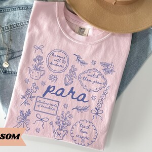 May include: A light pink t-shirt featuring a dark blue graphic print with the word "para" in the center, surrounded by illustrations of flowers, watering cans, bows, and text bubbles containing the phrases "learning with kindness," "watch them grow," "planting seeds of knowledge," and "teach love inspire."