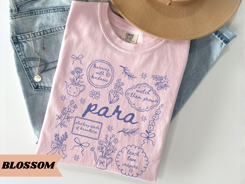 May include: A light pink t-shirt featuring a dark blue graphic print with the word "para" in the center, surrounded by illustrations of flowers, watering cans, bows, and text bubbles containing the phrases "learning with kindness," "watch them grow," "planting seeds of knowledge," and "teach love inspire."
