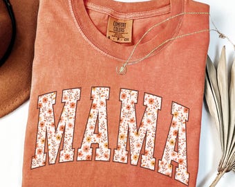 May include: A coral-colored Comfort Colors t-shirt with the word "MAMA" in large, floral-filled letters. A gold necklace with a pendant rests on the shirt. A brown hat and dried palm leaves are in the background.
