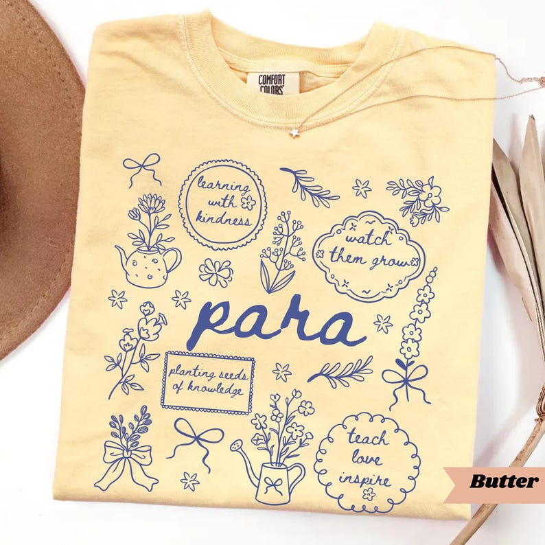 May include: A pale yellow t-shirt featuring dark blue line art illustrations of flowers, bows, and watering cans. The text reads: learning with kindness, watch them grow, para, planting seeds of knowledge, and teach love inspire.