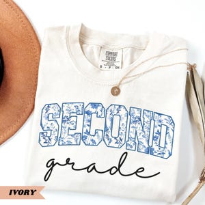 May include: Ivory t-shirt with a blue floral print that says "Second Grade" in a vintage style font.