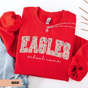 May include: A red crewneck sweatshirt with the word "EAGLES" in a floral design. The words "school name" are written in cursive below the word "EAGLES". A silver necklace with a pendant is on the sweatshirt. The word "RED" is at the bottom.