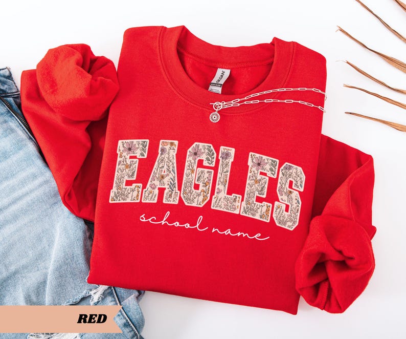 May include: A red crewneck sweatshirt with the word "EAGLES" in a floral design. The words "school name" are written in cursive below the word "EAGLES". A silver necklace with a pendant is on the sweatshirt. The word "RED" is at the bottom.