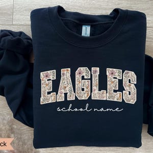 May include: Black crewneck sweatshirt with the word "EAGLES" in a floral design, with the words "school name" in a script font below. The sweatshirt is a solid black color.