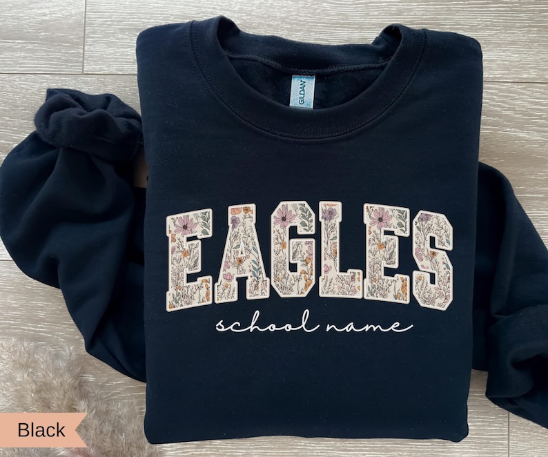 May include: Black crewneck sweatshirt with the word "EAGLES" in a floral design, with the words "school name" in a script font below. The sweatshirt is a solid black color.
