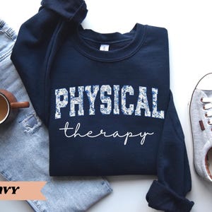 May include: Navy blue sweatshirt with the words "PHYSICAL therapy" in white script and floral print. The sweatshirt is paired with blue jeans, a coffee mug, and gray sneakers.