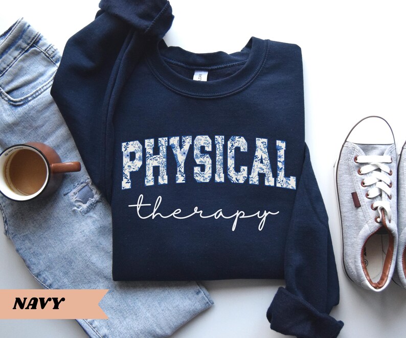 May include: Navy blue sweatshirt with the words "PHYSICAL therapy" in white script and floral print. The sweatshirt is paired with blue jeans, a coffee mug, and gray sneakers.