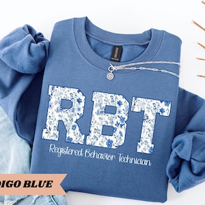 May include: Indigo blue sweatshirt with a white floral design that spells out "RBT" and the text "Registered Behavior Technician".