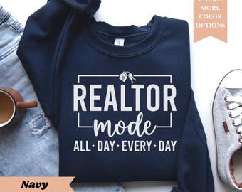 May include: Navy blue sweatshirt with the text "REALTOR mode ALL DAY EVERY DAY" in white. The design includes a house and key graphic. The sweatshirt is unisex and available in more color options.