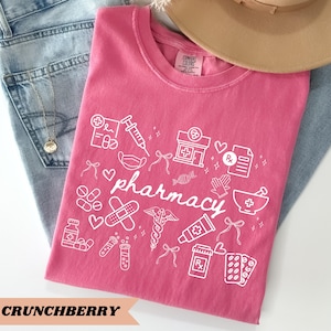 May include: A pink t-shirt with the word "pharmacy" and white pharmacy-related graphics. The design includes a syringe, pills, a pharmacy building, and a mortar and pestle. The shirt is folded on top of blue jeans.