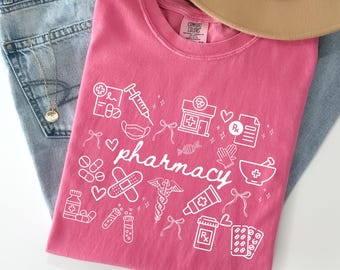Op de afbeelding: Een roze t-shirt met het woord "pharmacy" en witte apotheekgerelateerde afbeeldingen. Het ontwerp bevat een spuit, pillen, een apotheekgebouw en een vijzel en stamper. Het shirt ligt op een blauwe spijkerbroek.