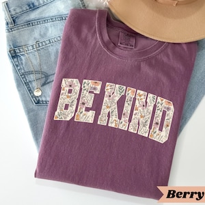 May include: A berry-colored t-shirt with the words "BE KIND" in a floral design. The shirt is folded on top of a pair of blue jeans and a tan hat. A gold necklace is also visible.