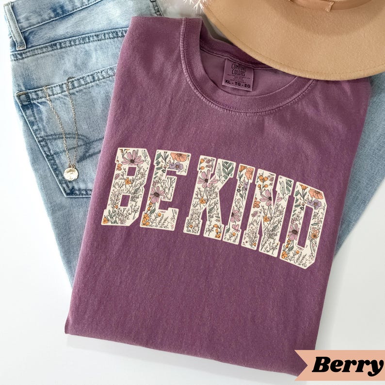 May include: A berry-colored t-shirt with the words "BE KIND" in a floral design. The shirt is folded on top of a pair of blue jeans and a tan hat. A gold necklace is also visible.