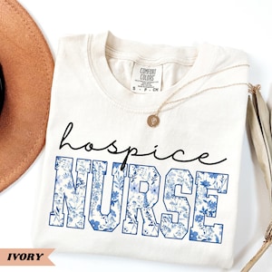 May include: A white t-shirt with the text "hospice NURSE" in blue lettering with a floral pattern. The shirt is on a white surface with a brown hat and a gold necklace.