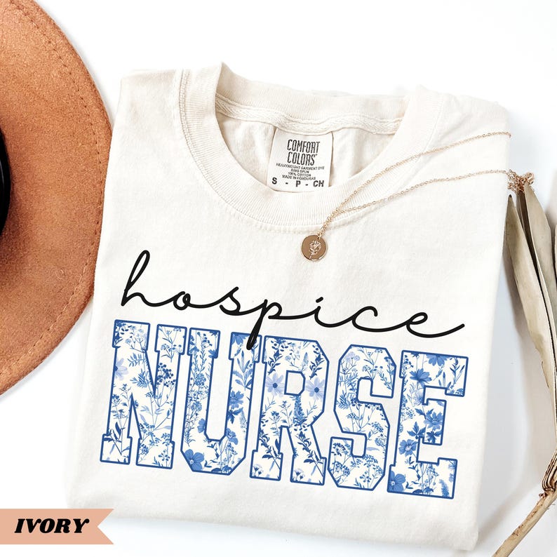 May include: A white t-shirt with the text "hospice NURSE" in blue lettering with a floral pattern. The shirt is on a white surface with a brown hat and a gold necklace.