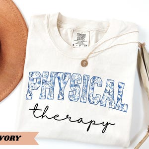 May include: Ivory-colored t-shirt with the words "PHYSICAL" and "therapy" printed on the front. The word "PHYSICAL" is in blue block letters filled with a floral pattern. A gold necklace rests on the shirt.