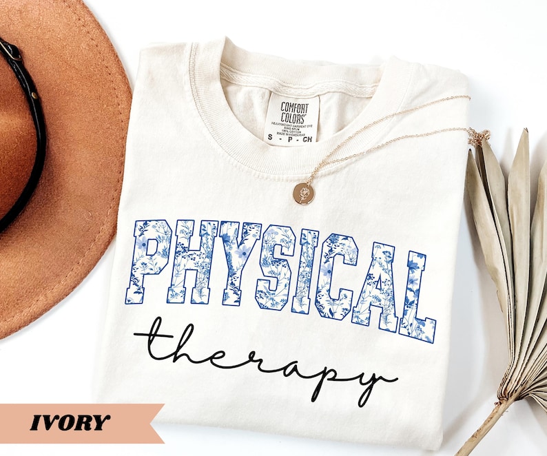 May include: Ivory-colored t-shirt with the words "PHYSICAL" and "therapy" printed on the front. The word "PHYSICAL" is in blue block letters filled with a floral pattern. A gold necklace rests on the shirt.