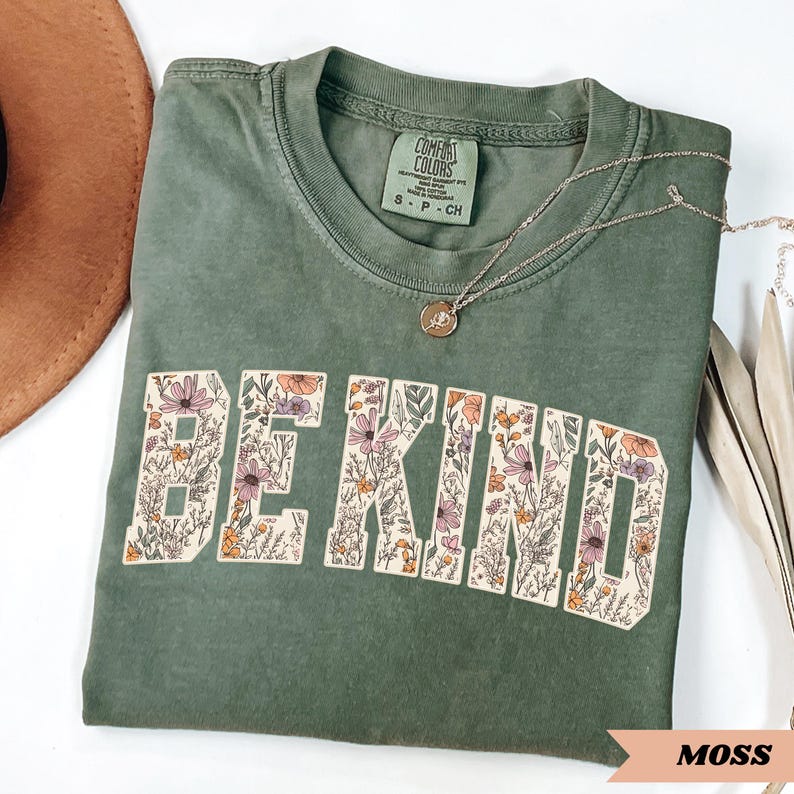 May include: A moss green t-shirt with the words "BE KIND" in a floral design. The shirt has a crew neck and short sleeves. A silver necklace with a pendant is draped across the shirt. The shirt is folded on a white surface.