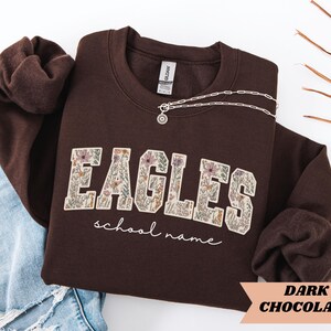 May include: A dark chocolate-colored sweatshirt with the word "EAGLES" in a floral design. The words "school name" are written in a script font below the word "EAGLES". A silver necklace with a pendant is draped across the sweatshirt.