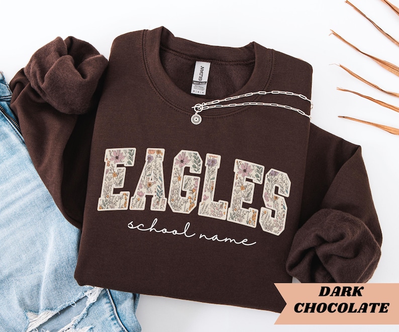 May include: A dark chocolate-colored sweatshirt with the word "EAGLES" in a floral design. The words "school name" are written in a script font below the word "EAGLES". A silver necklace with a pendant is draped across the sweatshirt.