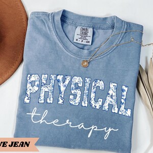 May include: A blue jean colored t-shirt with the words "PHYSICAL therapy" printed on the front. The word "PHYSICAL" is in a white font with a blue floral design. A gold necklace with a pendant is draped on the shirt.