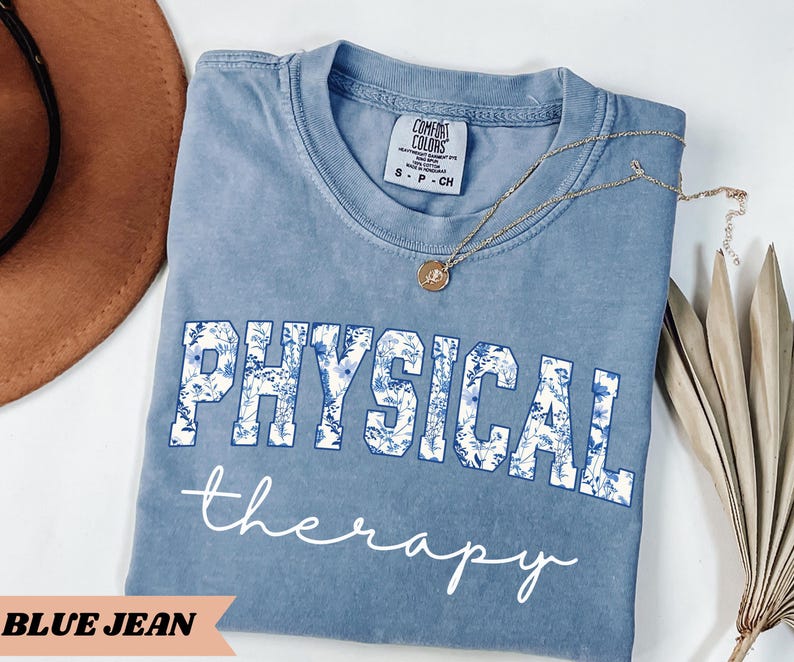 May include: A blue jean colored t-shirt with the words "PHYSICAL therapy" printed on the front. The word "PHYSICAL" is in a white font with a blue floral design. A gold necklace with a pendant is draped on the shirt.